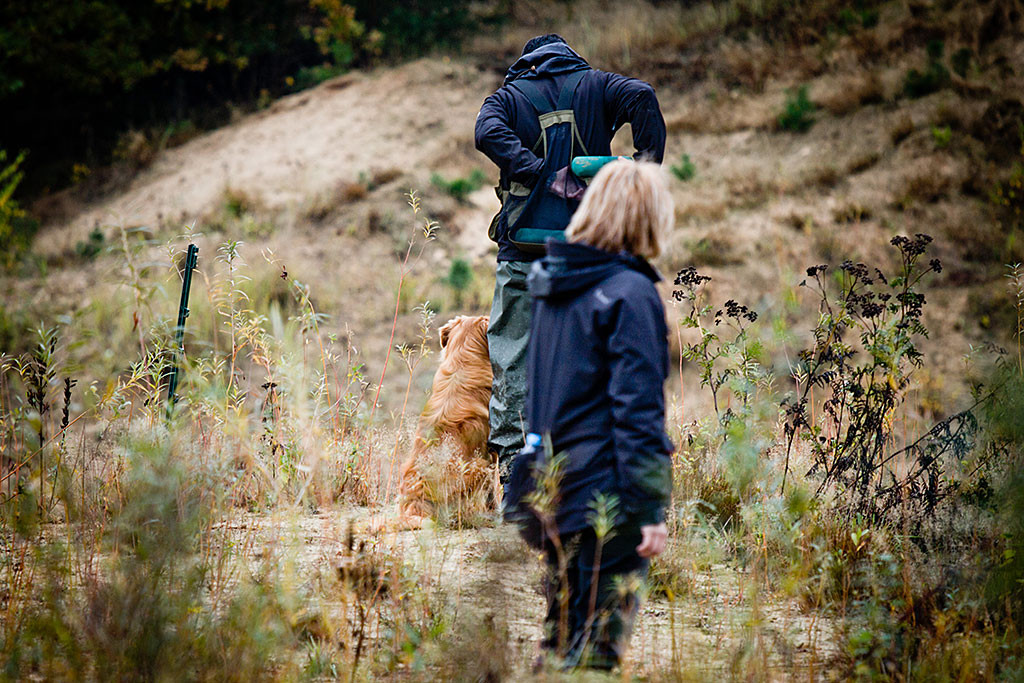 Edison sitzt neben Stephan, der gerade den zurückgebrachten Dummy in seine Weste steckt, im Vordergrund sieht man eine Helferin