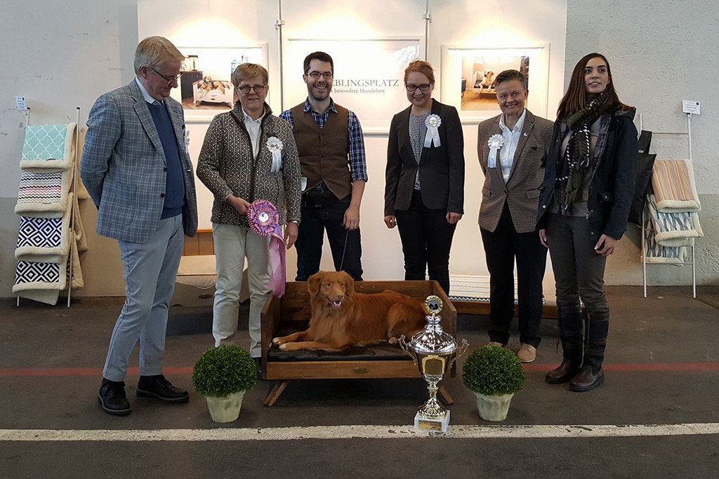 Edison liegt in einem Hundebett, vor ihm steht der Siegerpokal, hinter ihm stehen Stephan sowie alle vier Richter und die Sponsorin des Hundemöbels